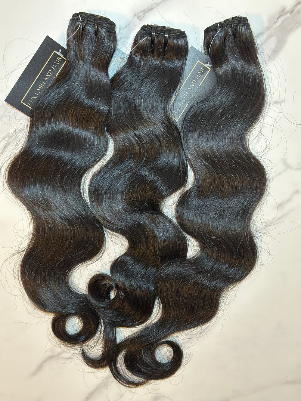 Three bundles of 100% raw Cambodian natural wave hair extensions from Lux Lash and Hair, with a soft, flowing wave pattern and natural shine, displayed on a marble background.