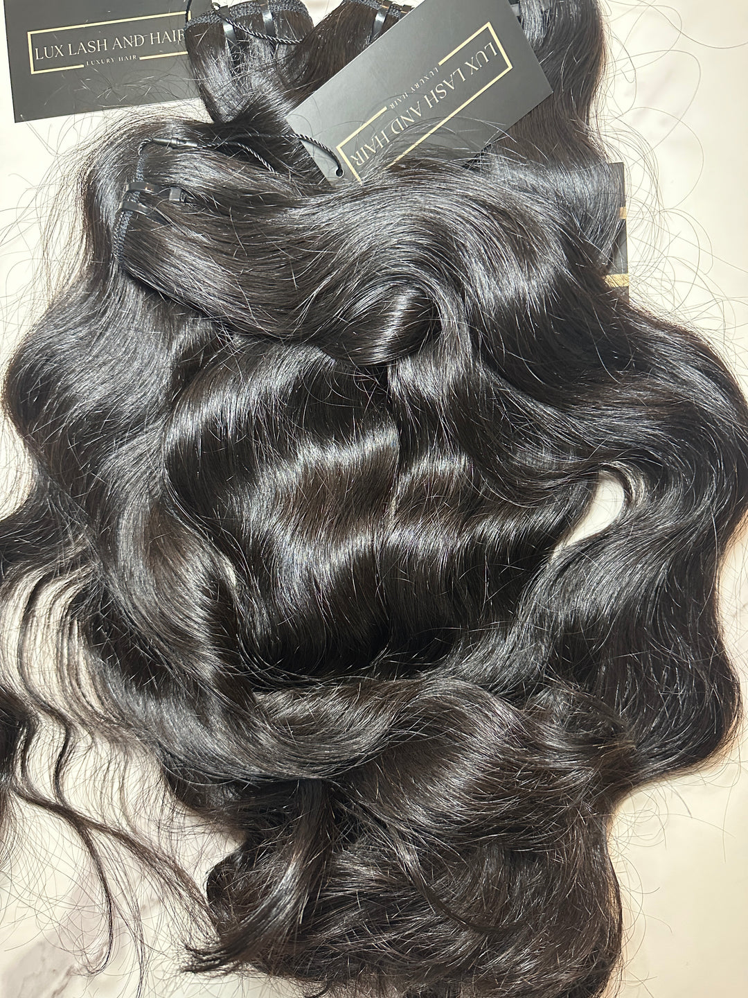 Four bundles of natural wave raw human hair extensions from Lux Lash and Hair, displayed on a light marble surface. The hair is dark, shiny, and full from root to tip, with soft, defined waves and attached black and gold brand tags.