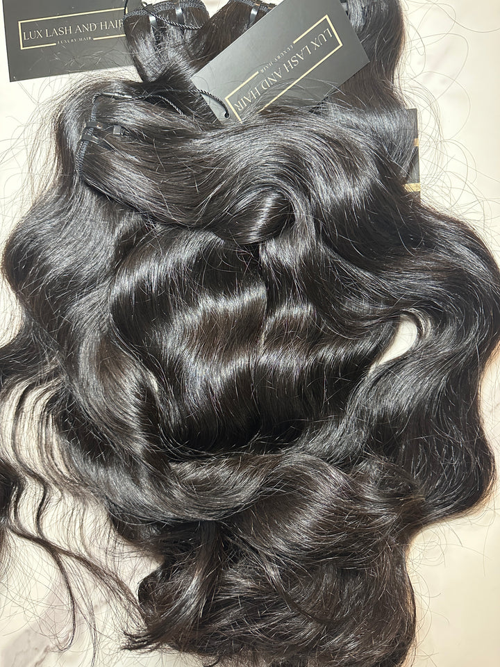 Four bundles of natural wave raw human hair extensions from Lux Lash and Hair, displayed on a light marble surface. The hair is dark, shiny, and full from root to tip, with soft, defined waves and attached black and gold brand tags.